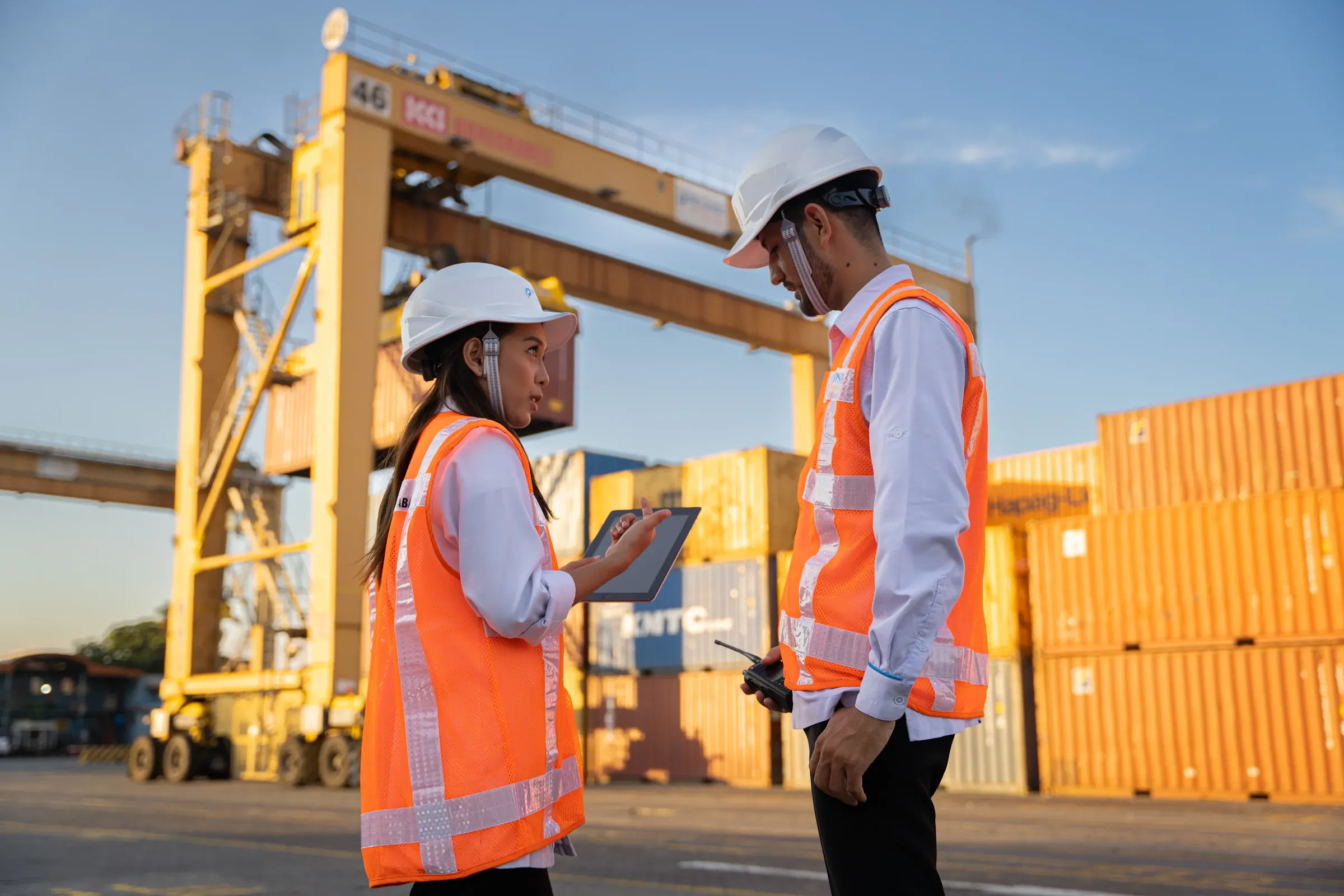 Port staff in high-visibility vests reviewing shipment documents on a tablet near stacked containers